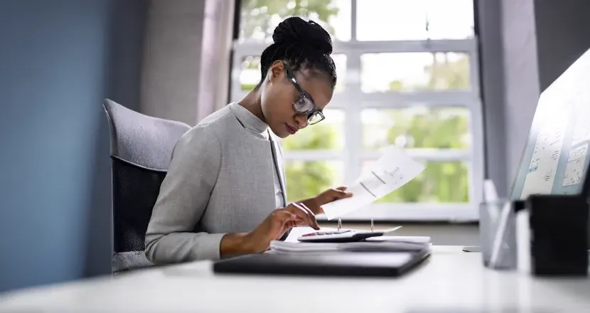 A Herzing University accounting student in professional attire reviews financial documents at a modern office desk, preparing reports and analyzing data.